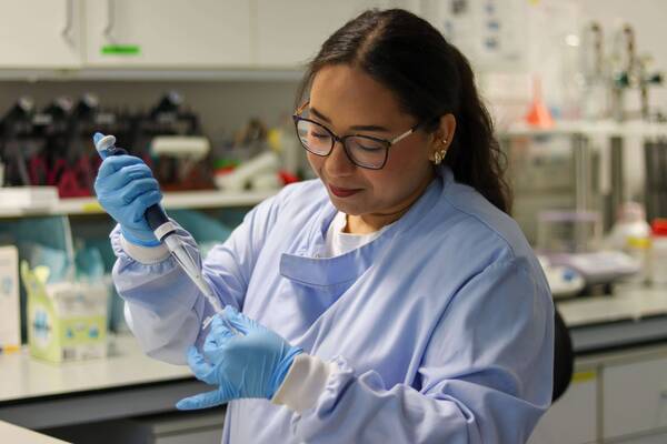 A focused scientist in a blue lab coat and gloves attaches a tip to a pipette in a lab.